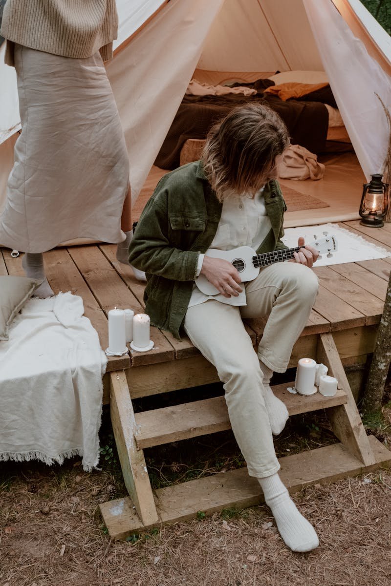 A man sits on outdoor steps playing ukulele beside a cozy tent with candles.